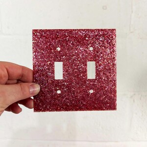 May include: A red glitter-covered light switch plate with two switch apertures and screw holes. The square plate is held up against a white wall. The finish is sparkly and decorative.