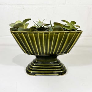 May include: A dark olive green ceramic planter with a fluted, rectangular bowl shape resting on a stepped pedestal base. The planter contains several small, light green artificial succulent plants with rounded leaves arranged inside the bowl.