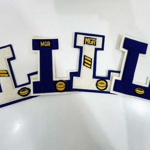 May include: Four embroidered letter patches shaped like the letter "L". Each patch has a navy blue border and a white centre. The top of each patch has the letters "MGR" in navy blue. Each patch features a football graphic.