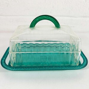 May include: A clear glass butter dish with a green plastic lid and handle. The dish has a decorative, wavy pattern.