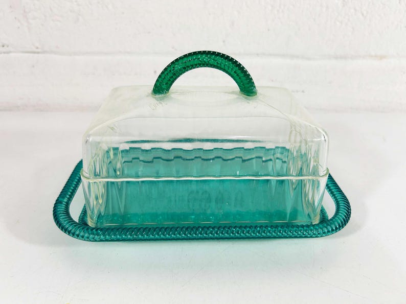 May include: A clear glass butter dish with a green plastic lid and handle. The dish has a decorative, wavy pattern.