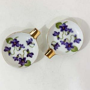 May include: Two small, white ceramic ashtrays with a gold rim and handle. Each ashtray is decorated with a floral pattern of purple violets and green leaves. The ashtrays are likely vintage.