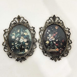 Puede incluir: Dos marcos de pared ovalados con bordes ornamentados de volutas metálicas oscuras. Cada marco contiene una impresión de un arreglo floral con varias flores coloridas sobre un fondo oscuro, dentro de una carcasa de metal estilo filigrana.
