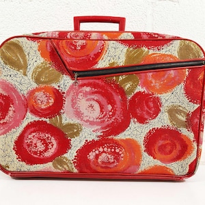 Könnte beinhalten: Ein Vintage-Koffer mit einem lebendigen Blumenmuster in Rot-, Orange- und Rosatönen. Der Koffer hat einen roten Griff, Zierleisten und einen schwarzen Reißverschluss. Das Design umfasst große, stilisierte rosenartige Formen auf cremefarbenem Hintergrund.