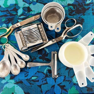 Puede incluir: Una colección de utensilios de cocina vintage, que incluye cucharas medidoras, tijeras, un cortador de queso y tazas medidoras. Las herramientas están hechas de metal y plástico, y están dispuestas sobre una superficie con estampado floral azul.