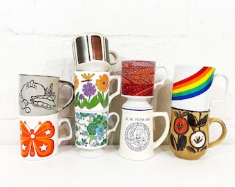 May include: A stack of vintage ceramic mugs with various designs. The mugs feature colorful patterns, including a butterfly, flowers, a rainbow, and abstract shapes. Some mugs have text, and the colors include orange, brown, and white.