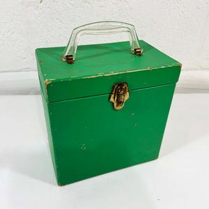 Pode incluir: Uma caixa de madeira verde vintage com uma alça de plástico transparente e um fecho de metal dourado. A caixa tem formato retangular com uma frente ligeiramente inclinada. A tinta mostra sinais de desgaste.
