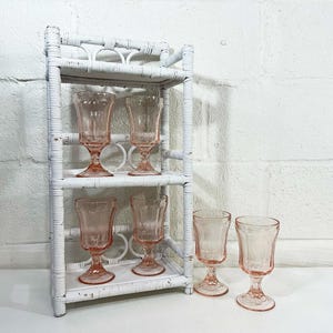 May include: A white wicker shelf unit displays six pink stemmed glasses. The glasses have a delicate, etched design. The shelf is against a white brick wall, creating a vintage aesthetic.