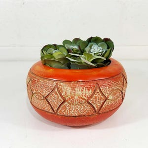 May include: A round, orange ceramic planter with a textured, cream-colored diamond pattern around the center. The planter contains several green artificial succulent plants with layered, fleshy leaves that fill the opening of the vessel.