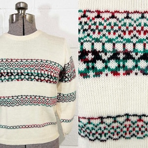 May include: Cream-colored sweater with a patterned design in red, green, and black. The sweater features a crew neck and long sleeves. The pattern is repeated on the sleeves and the body of the sweater.