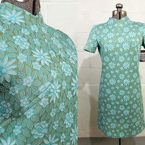 May include: A short-sleeved dress featuring a high mandarin collar and a textured floral pattern in shades of light blue and olive green. The garment is displayed on a dress form and includes a small tag attached to the side of the sleeve.