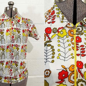May include: A short-sleeved shirt with a white background and a repeating floral pattern in red, yellow, and orange. The shirt has a yellow zipper and a collar. The shirt is displayed on a mannequin.