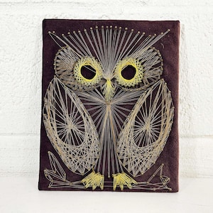May include: A rectangular wall art piece featuring a string art owl design. The owl is created with thin gold and yellow threads wrapped around metal nails on a dark brown, textured fabric background, forming a detailed geometric bird shape.