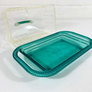 May include: A teal blue glass dish with a raised rim and a clear glass lid. The dish has a decorative pattern around the edge.