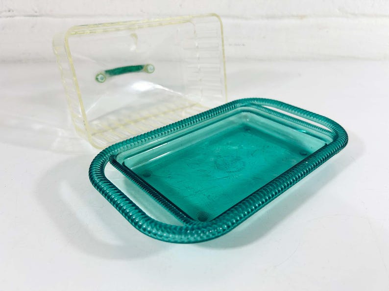May include: A teal blue glass dish with a raised rim and a clear glass lid. The dish has a decorative pattern around the edge.
