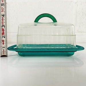 May include: A clear plastic butter dish with a green lid and base. The lid has a handle and the base has a decorative edge.