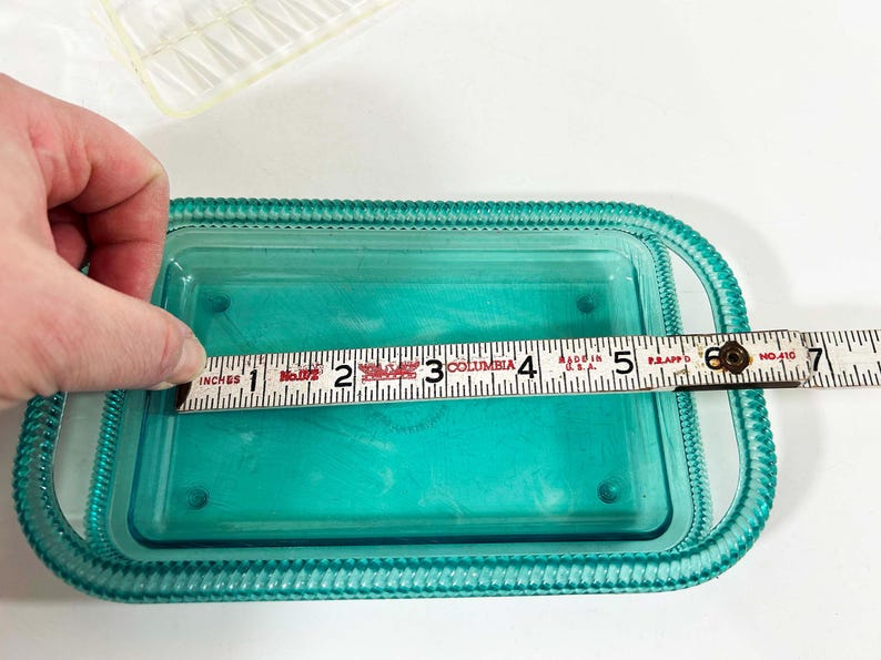 May include: A teal blue glass dish with a raised rim and a beaded edge. The dish is 5 inches long and is being measured with a ruler.