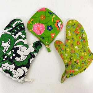 May include: Two quilted oven mitts and one square pot holder. One mitt features a bold black, white, and green paisley pattern. The other mitt and the square pot holder display a vibrant green fabric with a colorful floral and small nature-themed print.