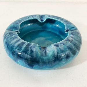 May include: A round, ceramic ashtray with a glossy, turquoise and blue glaze. The ashtray has a wide rim with three notches for resting cigarettes. The interior is a solid turquoise color.