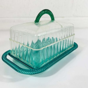 May include: A clear plastic butter dish with a green lid and handle. The dish has a ribbed design and a green plastic base.