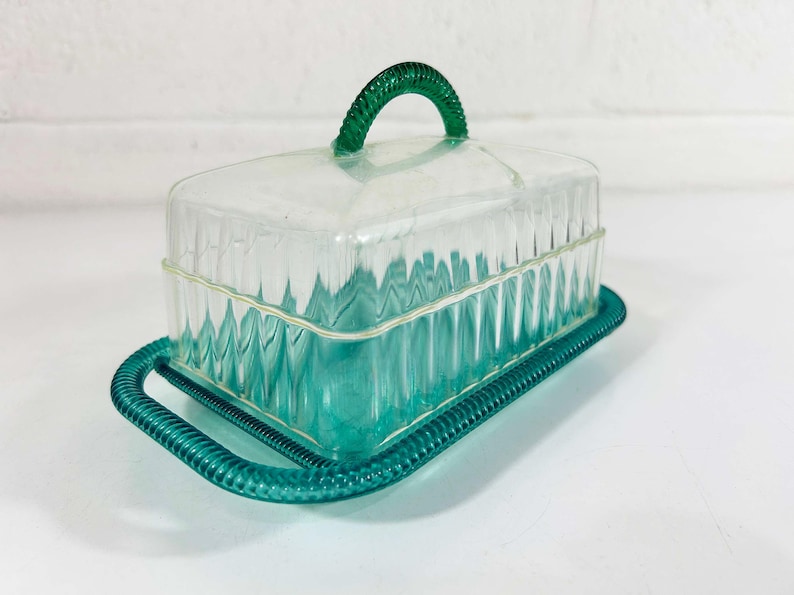 May include: A clear plastic butter dish with a green lid and handle. The dish has a ribbed design and a green plastic base.