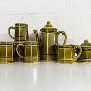 May include: A complete olive green ceramic tea set, including a teapot, sugar bowl, creamer, and four mugs. The set has a geometric design with vertical and horizontal lines. The teapot has a lid and a curved handle. The mugs have a curved handle.