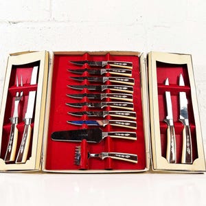 Op de afbeelding: Een vintage bestekset in een crèmekleurige doos met een rode binnenkant. De set bevat steakmessen, trancheermessen, vorken en een vleesvermalser. De handvatten hebben een crème- en bruin ontwerp.