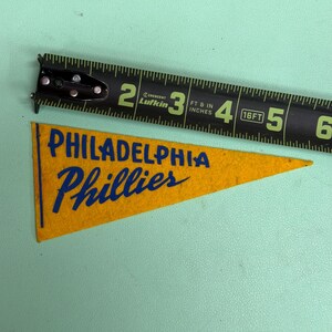 May include: A vintage Philadelphia Phillies pennant in yellow felt with blue lettering. A black tape measure shows the pennant is approximately 4 inches long. The pennant is on a light green surface.