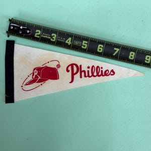 May include: Cream-colored pennant with the word "Phillies" in red script, along with a red baseball and cap graphic. A black tape measure is next to the pennant. The pennant has a black border on the left side.