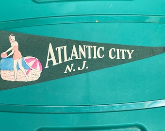May include: A green felt pennant with white text that reads "ATLANTIC CITY N.J."  The pennant features a cartoon illustration of a woman in a swimsuit holding a beach ball and an umbrella.