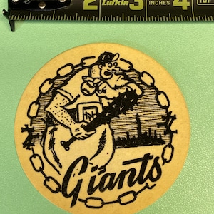 May include: A vintage, cream-colored, round paper coaster with a black and white illustration of a baseball player holding a bat. The player is wearing a baseball uniform with the letters "NY" on the chest. The text "Giants" is written in a cursive font below the player.