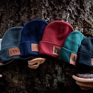 May include: A collection of six beanies in various colors, including gray, navy, red, and teal. Each beanie features a leather patch with outdoor-themed designs like mountains, a bear, and a tree. The beanies are displayed on a tree trunk.