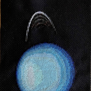 Puede incluir: Un punto de cruz azul y blanco de un planeta con un anillo a su alrededor. El planeta está rodeado de un fondo negro.