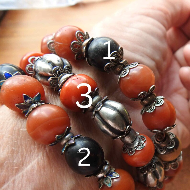 May include: Three bracelets with orange and black beads. The beads are separated by silver spacers with intricate floral designs. The bracelets are numbered 1, 2, and 3.
