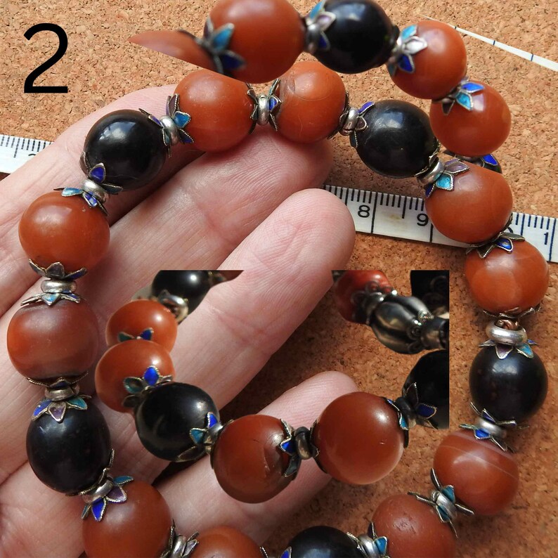 May include: A beaded bracelet with alternating amber and black beads. Each bead has a silver accent with blue enamel. The bracelet is on a hand, and the number "2" is visible in the background.
