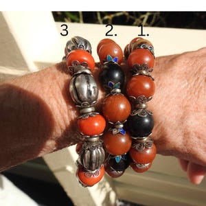 May include: Three bracelets with amber beads, silver accents, and black beads with blue enamel accents. The bracelets are stacked on a person's wrist.