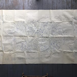 Könnte beinhalten: Ein großes, gefaltetes Blatt gealtertes Papier mit einer detaillierten blau-schwarzen Tuschezeichnung. Das Kunstwerk zeigt ein zentrales Blumendesign mit umliegenden Illustrationen und Text am unteren Rand. Das Papier hat Knicke und ein Vintage-Aussehen.