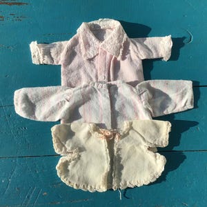 May include: Three vintage baby doll outfits. The top shirt is pink with a lace collar and cuffs. The middle shirt is pink with vertical stripes. The bottom is a cream-colored jacket with a scalloped edge and a small bow.