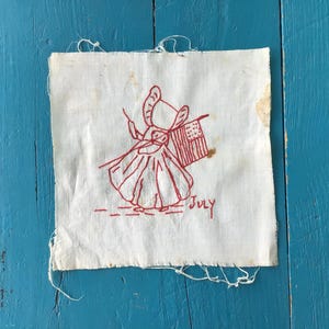 May include: A vintage white fabric square with a red embroidered design. The design features a figure holding a flag with stars and stripes, and the word "July" is embroidered below. The fabric has frayed edges and some discoloration.