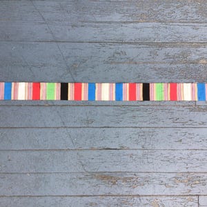 May include: A colorful, striped textile with a red fringe at each end. The stripes include red, blue, white, green, and black. The textile is lying on a weathered, gray wooden surface.