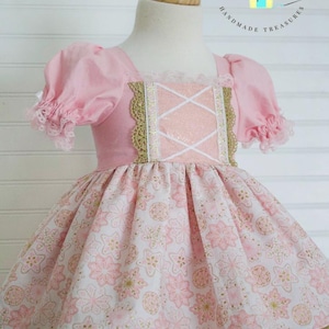 Puede incluir: Vestido rosa y blanco con estampado floral, ribetes dorados y detalles de encaje. El corpiño presenta un escote cuadrado y un diseño de cinta cruzada. El vestido tiene mangas cortas abullonadas y se exhibe en un maniquí.