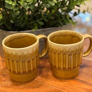 Könnte beinhalten: Zwei Keramikbecher in Braun- und Beigetönen mit einem gestapelten Design. Die Becher haben einen Henkel und ein geriffeltes Muster auf der unteren Hälfte. Sie stehen auf einer Holzoberfläche.