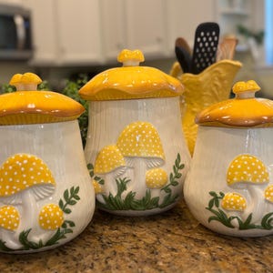 Puede incluir: Juego de tres botes de cerámica con forma de seta, con tapas amarillas y diseños de setas. Los botes son blancos con setas amarillas y blancas punteadas y hojas verdes. Las tapas son amarillas con ribete marrón.