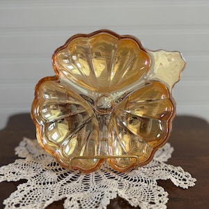 Puede incluir: Un plato de vidrio ámbar vintage con forma de flor de tres pétalos. El plato tiene un centro elevado y un borde festoneado. Está sentado sobre un encaje blanco de ganchillo.