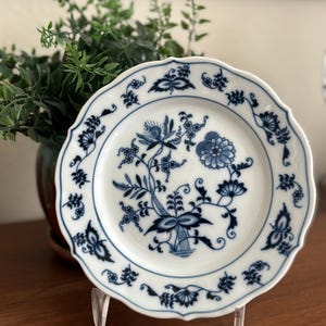 May include: White porcelain plate with a scalloped edge and a blue floral pattern. The plate features a central floral design and smaller floral motifs around the rim. The plate is displayed on a clear stand.
