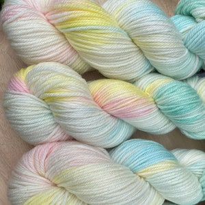 May include: Four skeins of yarn in a pastel colour palette. The yarn is a blend of white, yellow, pink, and blue. The yarn is arranged in a circle on a wooden surface. The yarn is labelled "Dreamy"
