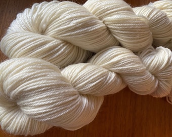 May include: Close-up of ivory-colored yarn skeins. The yarn is tightly wound, showing individual strands. The texture appears soft and suitable for knitting or crocheting projects. The yarn is displayed on a wooden surface.