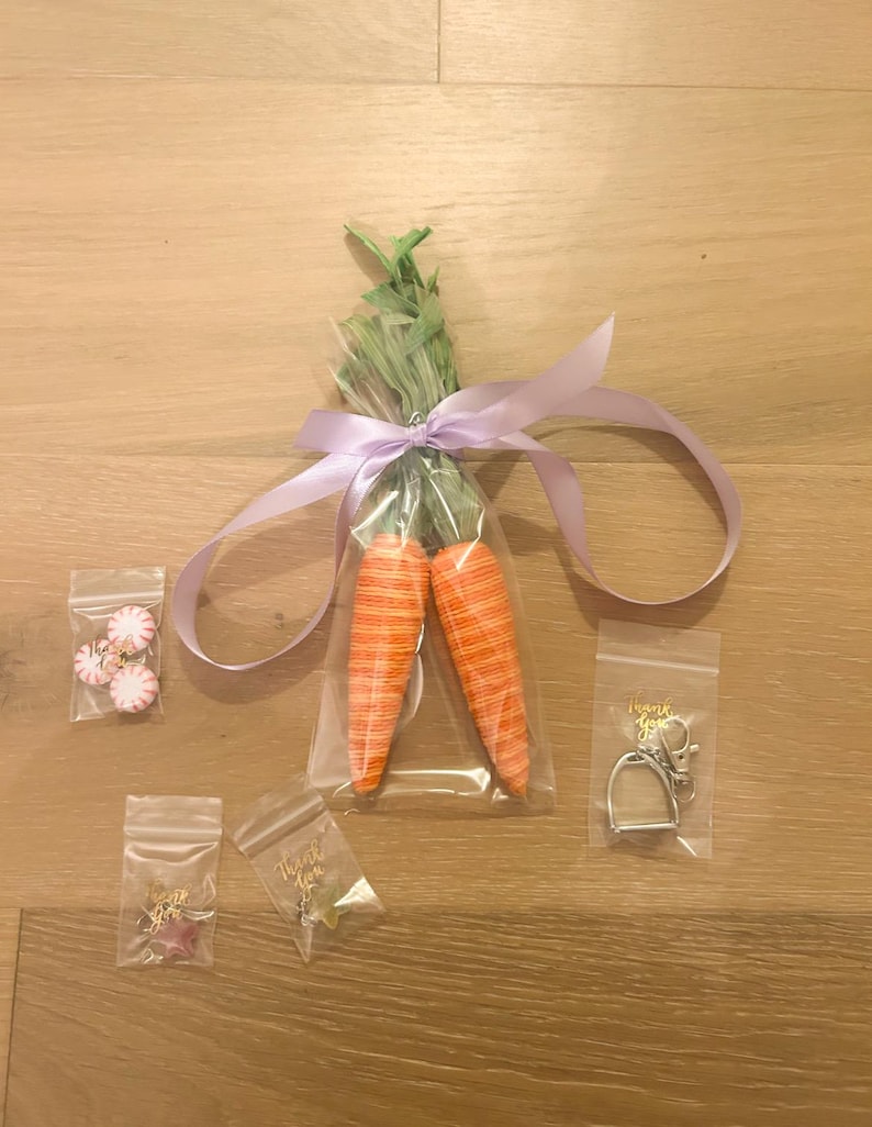 May include: Two orange and white carrot-shaped decorations tied together with a purple ribbon and packaged in a clear plastic bag.  There are four small bags with various small items, including a pair of earrings, a charm, and a key chain with a horseshoe charm.  The bags have the text "Thank You" printed on them.