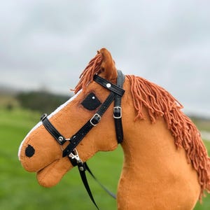 May include: A plush hobby horse toy featuring a tan fabric head with a white blaze on the nose, black felt eyes, and a textured yarn mane in a matching tan color. The horse is equipped with a black faux leather bridle with silver buckles.