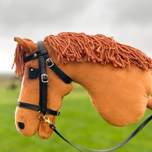 May include: A plush hobby horse with light brown fabric, a textured orange yarn mane, and a white blaze on the face. It features a black faux leather bridle with gold-toned metal buckles, a gold-toned clip, and long black reins attached to the side.